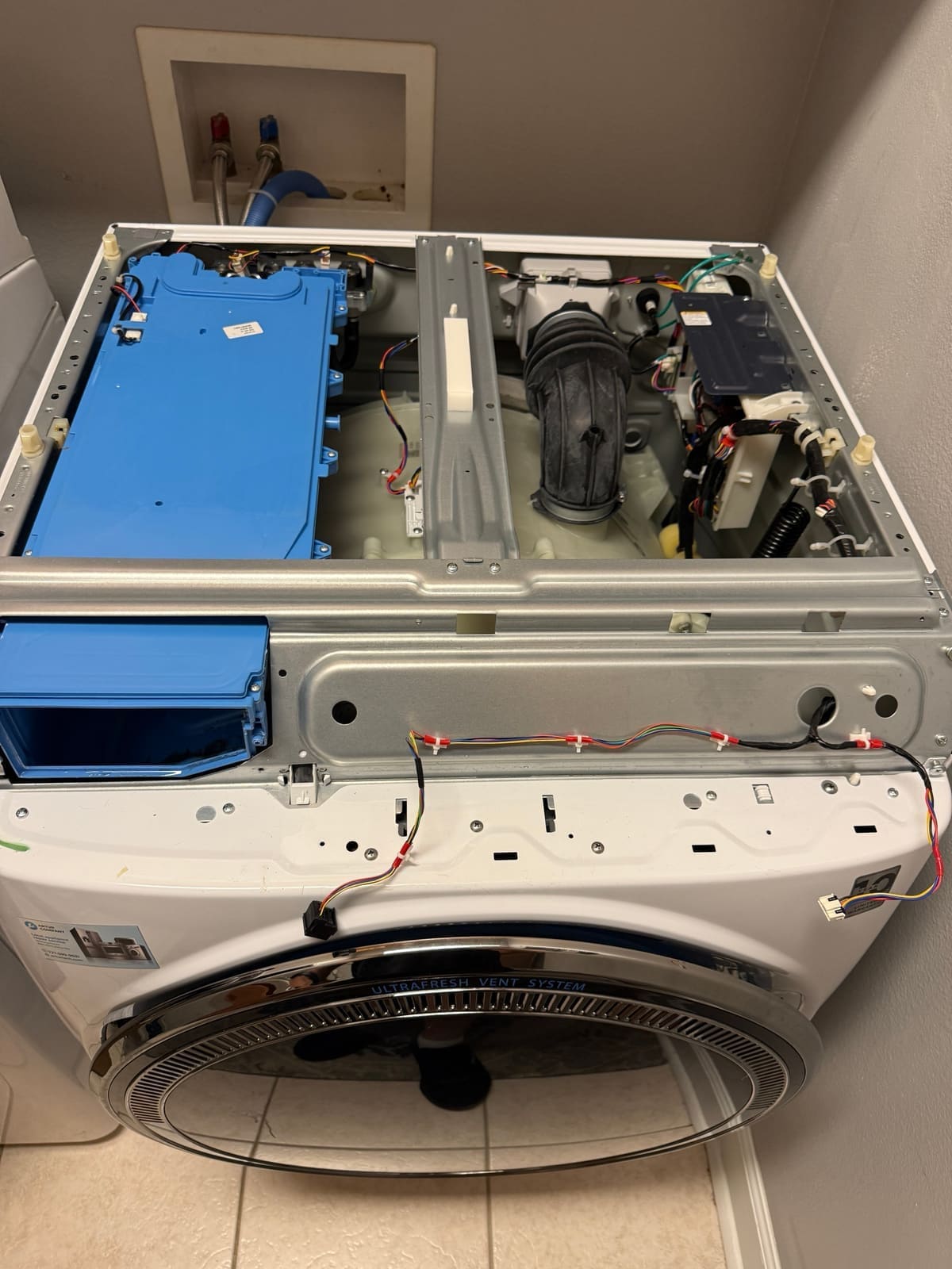 GE front-load washer with the top off, showing the blue dispenser assembly, drain hose and wiring