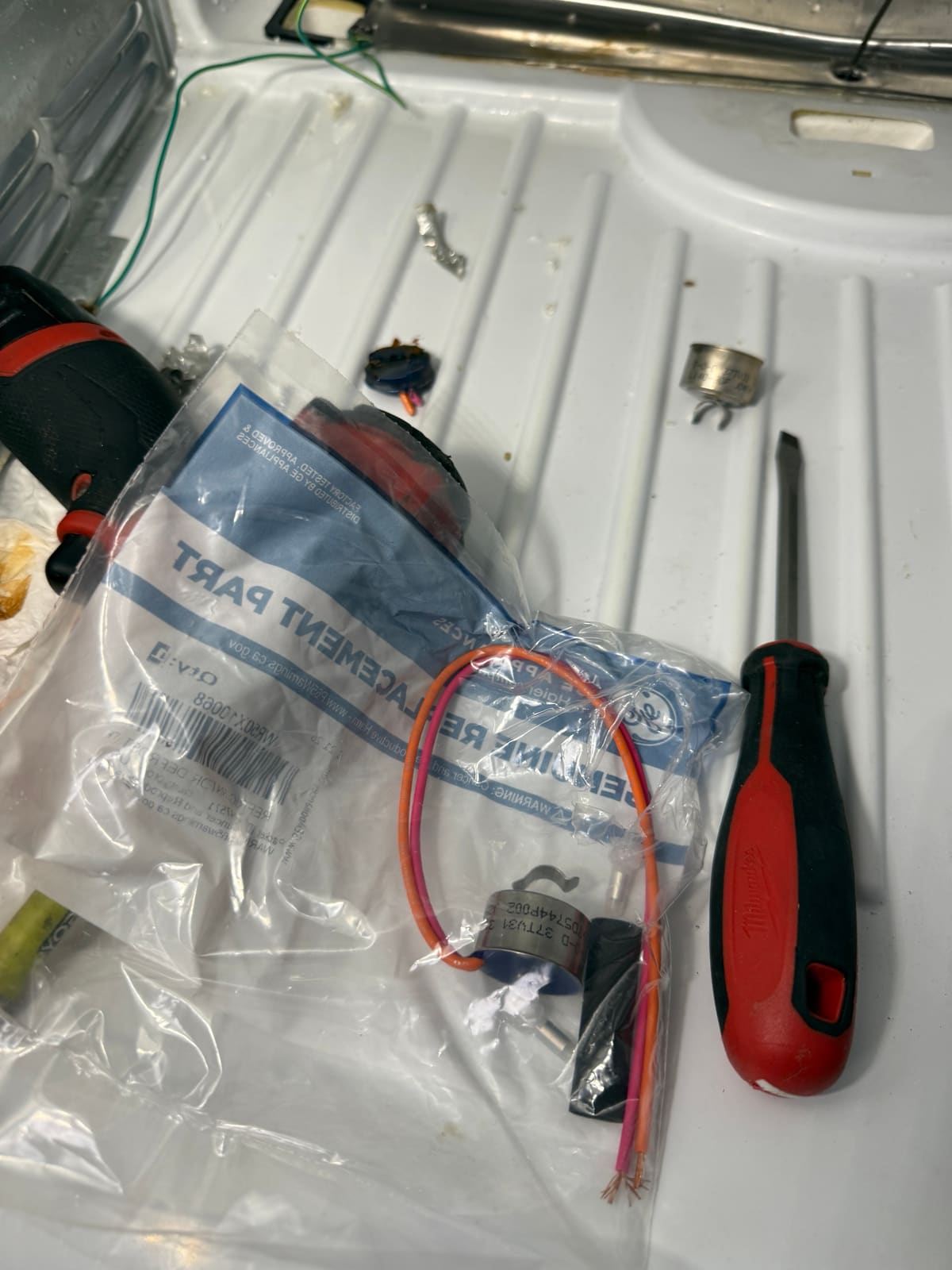 New GE defrost thermostat in its genuine replacement part bag, with the old thermostat nearby
