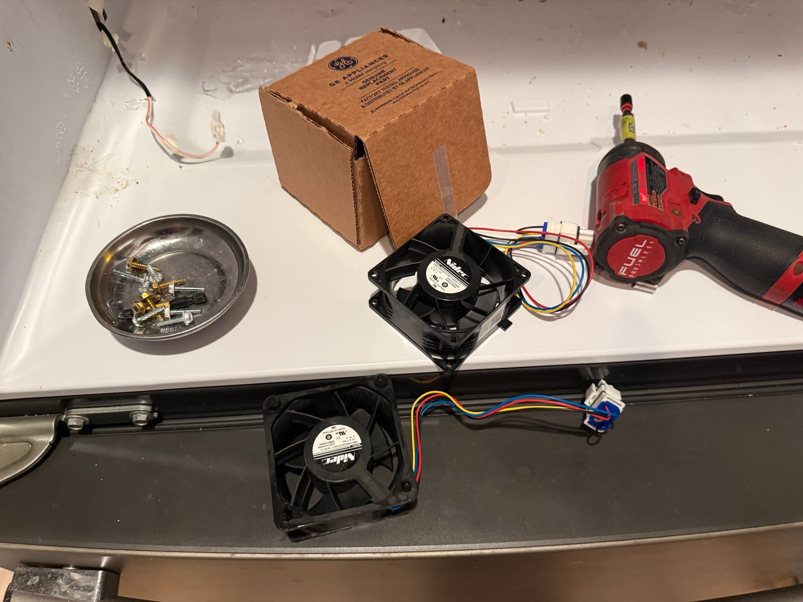 Old GE refrigerator fan and the new replacement fan side by side, with the GE parts box and a magnetic tray of screws