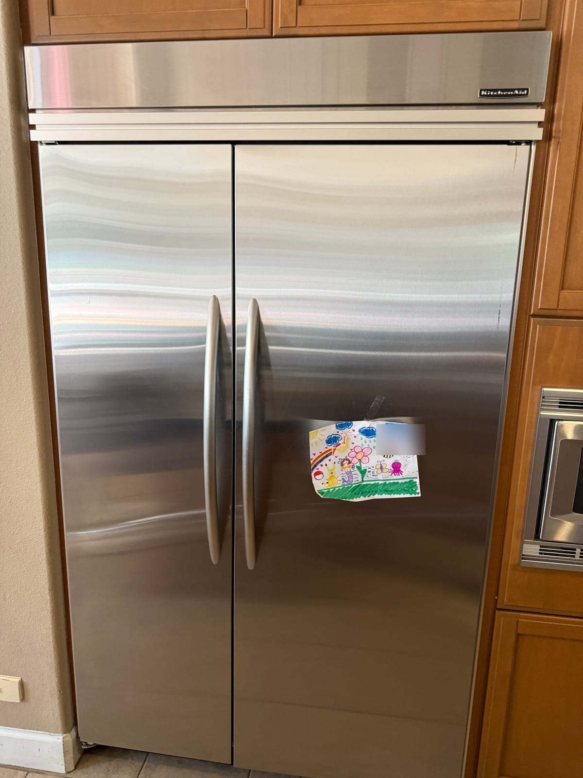 KitchenAid built-in side-by-side refrigerator with stainless doors and a child's drawing on the door, set into wood cabinetry in Palm Harbor
