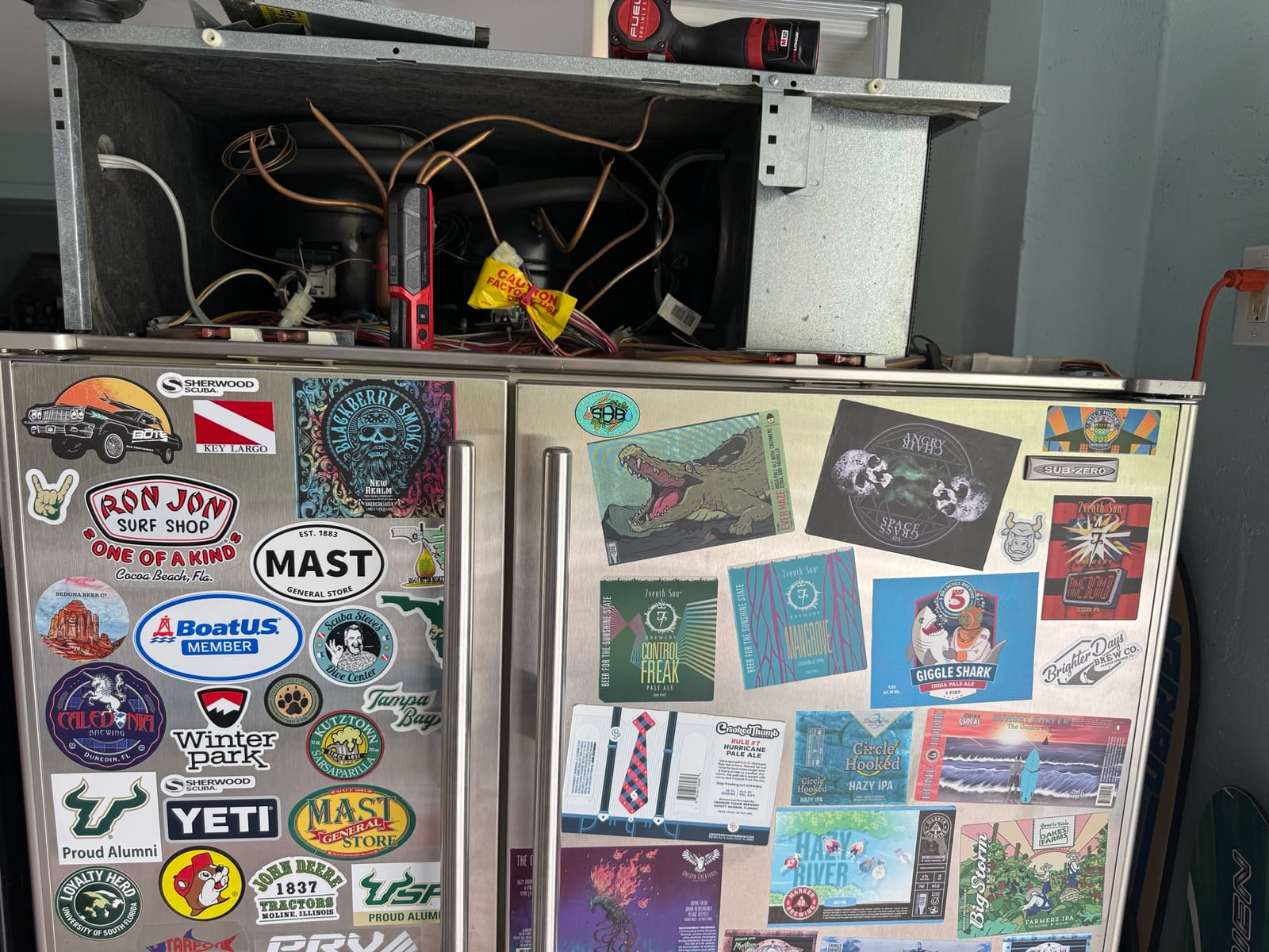 Built-in Sub-Zero side-by-side refrigerator covered in stickers, with the top grille off and the machine compartment open, in a garage in Tarpon Springs