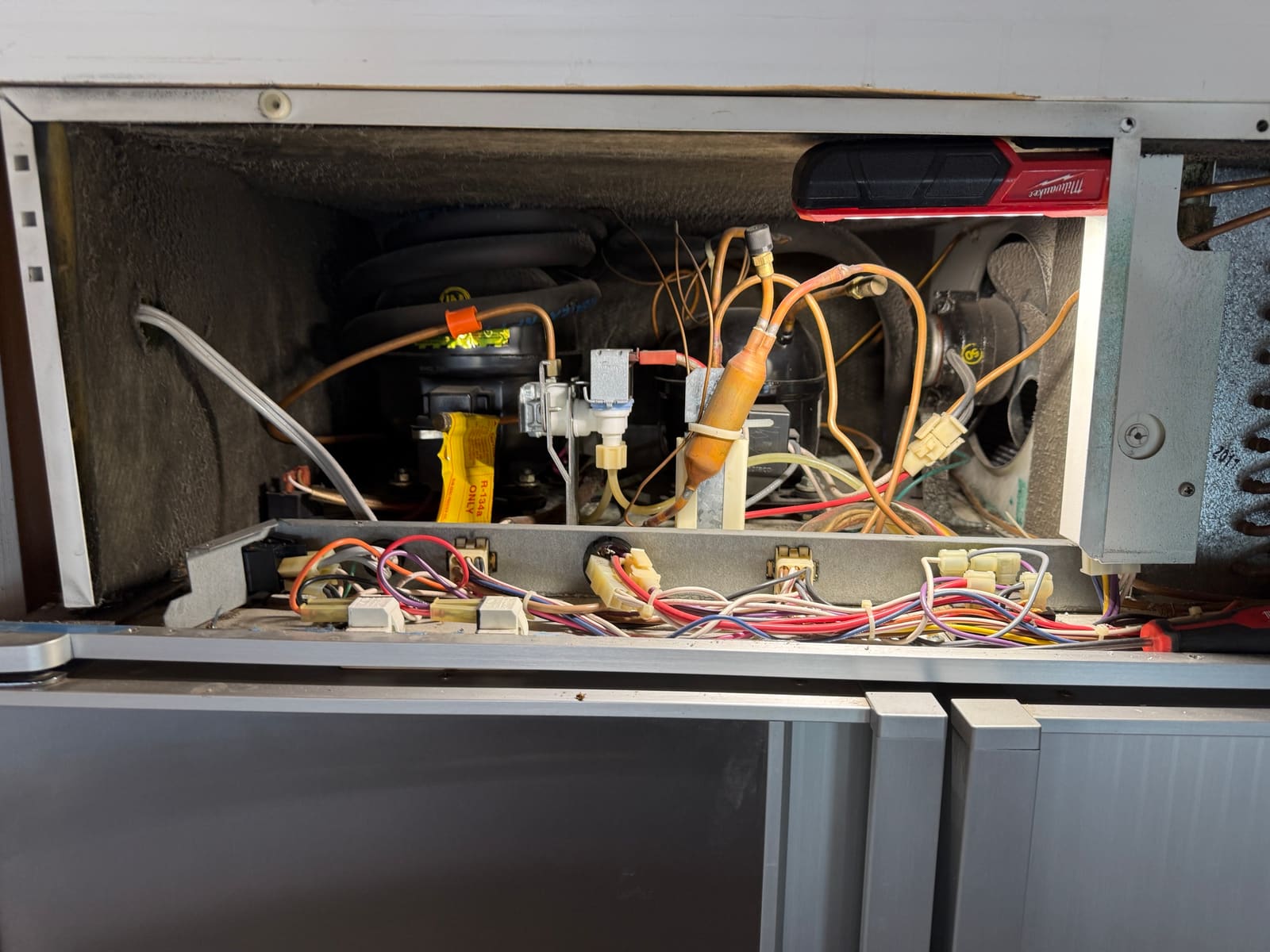 Open Sub-Zero machine compartment above the doors, showing the compressor, condenser fan, water valve and the wiring bracket along the front