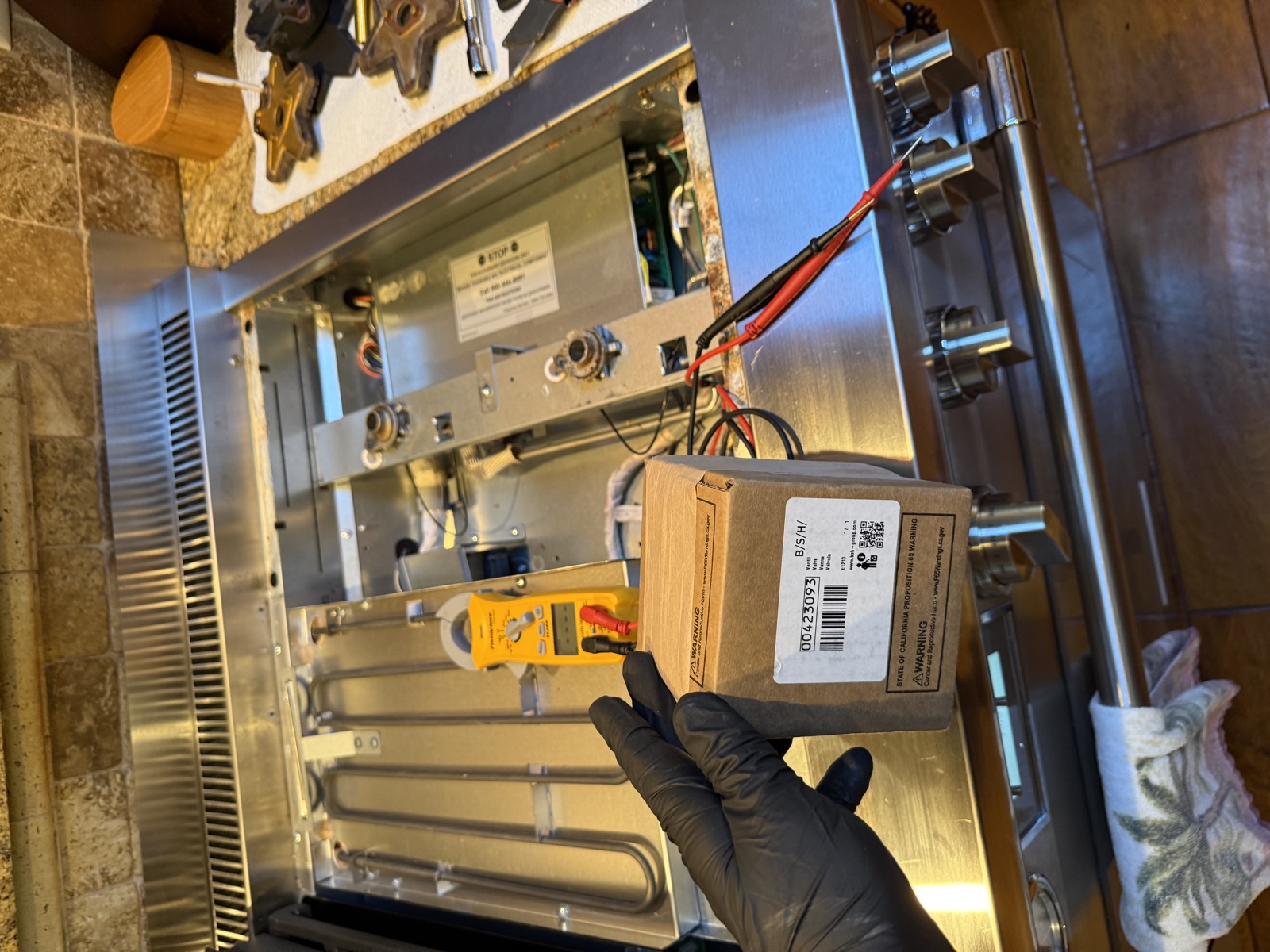 Boxed replacement burner valve held over the opened Thermador cooktop