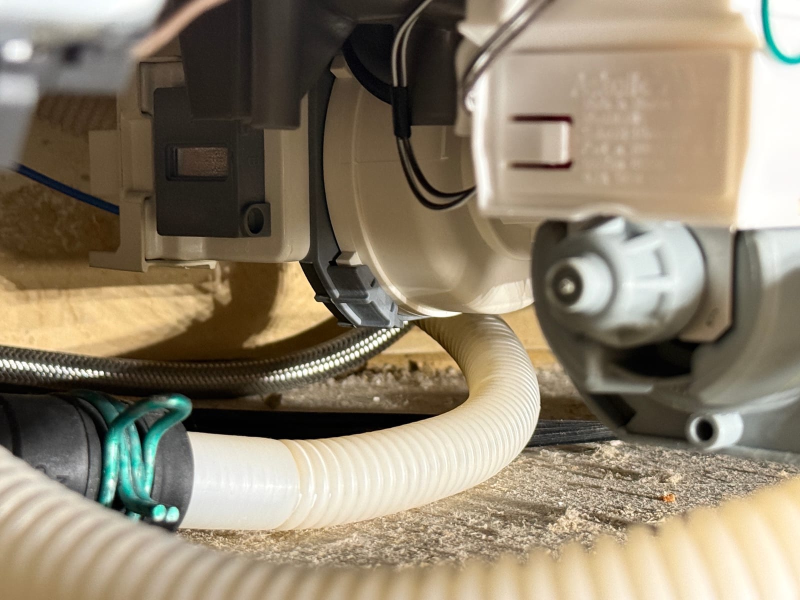 Close-up under the dishwasher showing the pump assembly, the corrugated drain hose and the braided water supply line