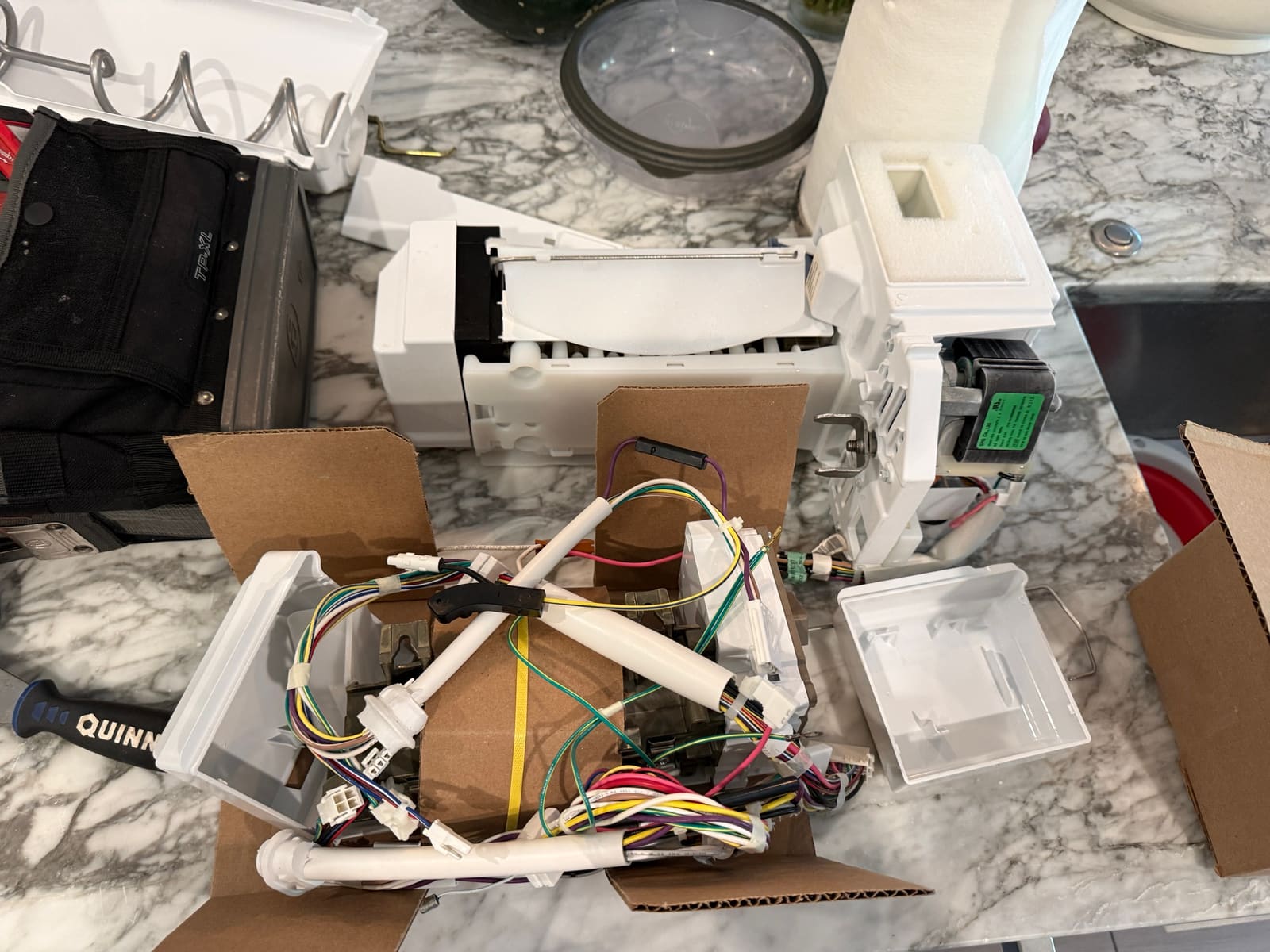 Old Whirlpool ice maker with its black electronic module next to the new ice maker kit unpacked from its box on a kitchen counter in Westchase