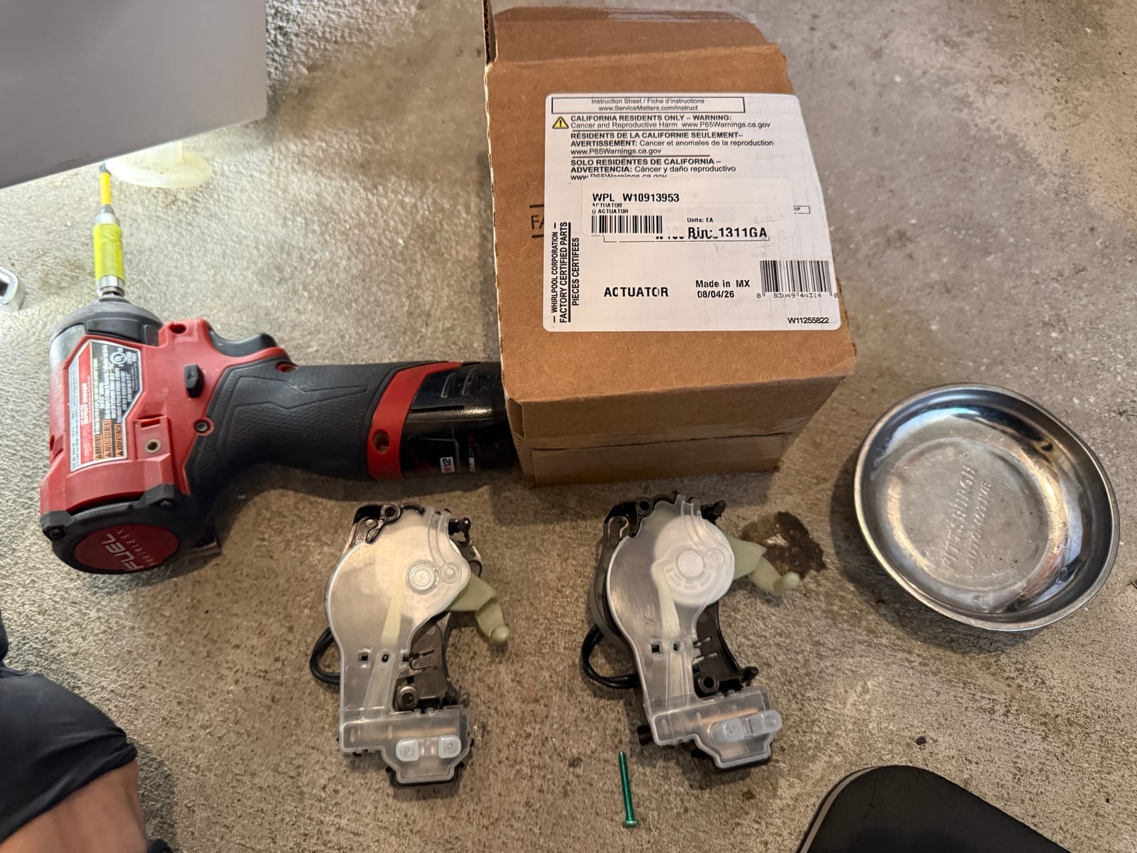 Two Whirlpool mode shifter actuators on the floor next to a genuine Whirlpool actuator box, with a drill and a parts tray beside them