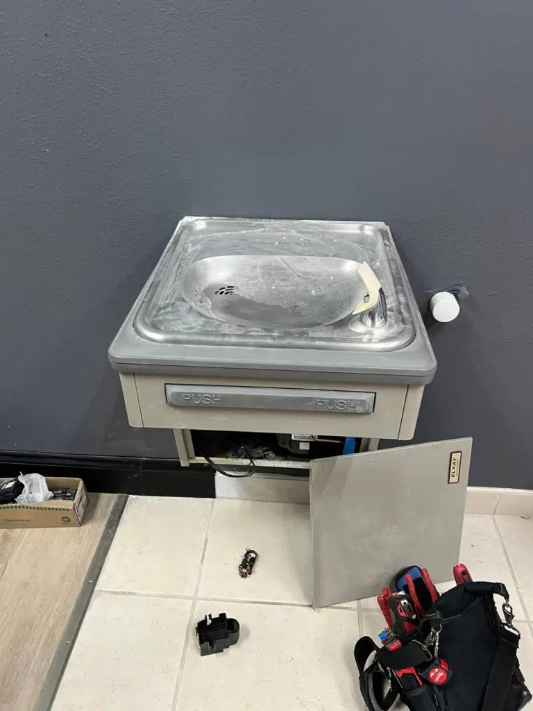 **Image Description:** A technician from Artur Appliance Repair is fixing a wall-mounted drinking fountain. The technician is using professional tools, ensuring the fountain's optimal performance. The background shows a clean and well-lit public space with the technician focused on the repair task. Various parts and tools are neatly arranged around the workspace. **Alt Text:** Technician from Artur Appliance Repair fixing a wall-mounted drinking fountain.